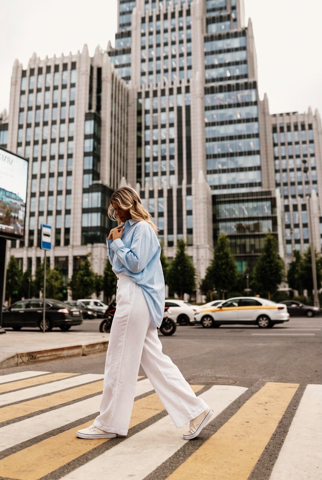 Customer in a blue striped oversized shirt and white wide-leg trousers crossing a city street